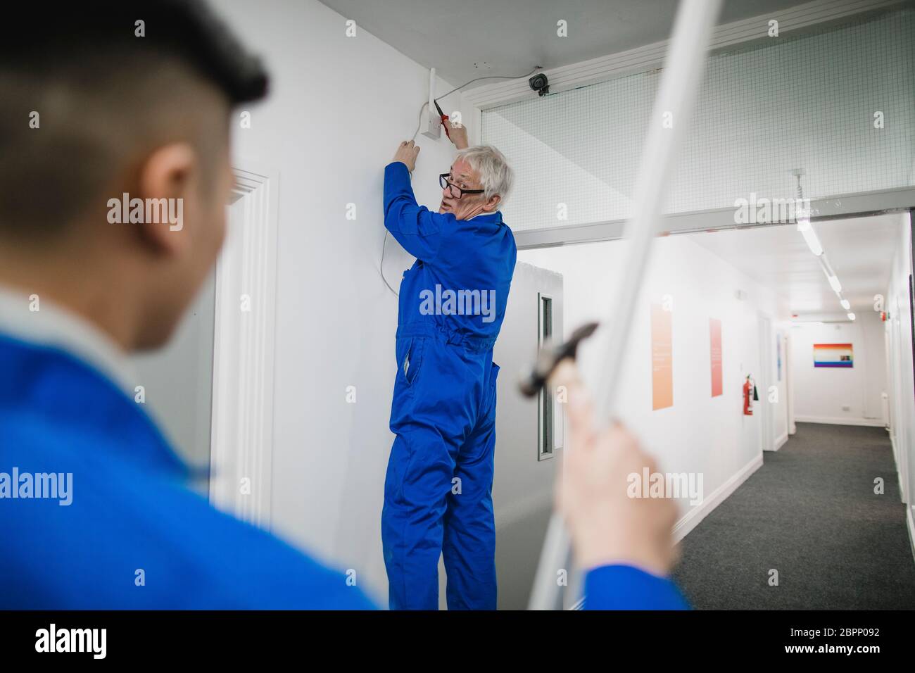 Senior electrician is talking to his trainee while fitting a security camera in the corridor of an office building. Stock Photo