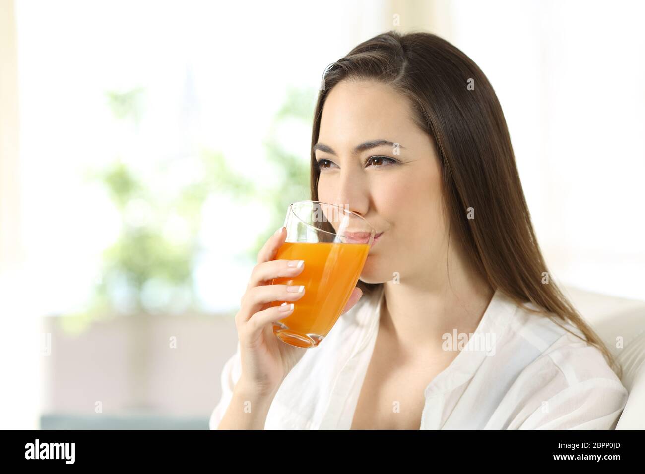 Woman drinking orange juice in a glass sitting on a couch in the living room at home Stock Photo