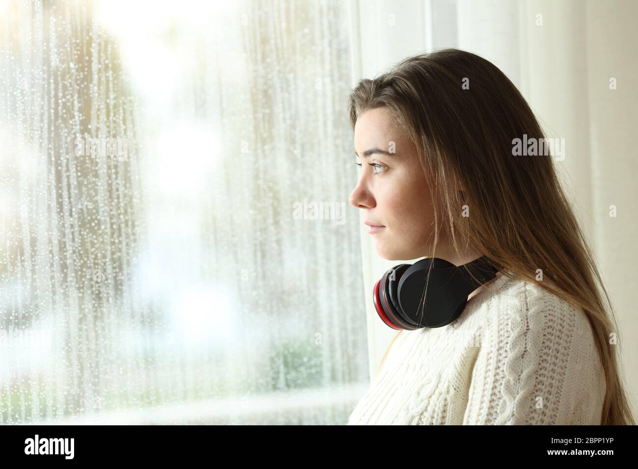 Side view portrait of a sad teen with headphones looking through a window in a rainy day Stock Photo