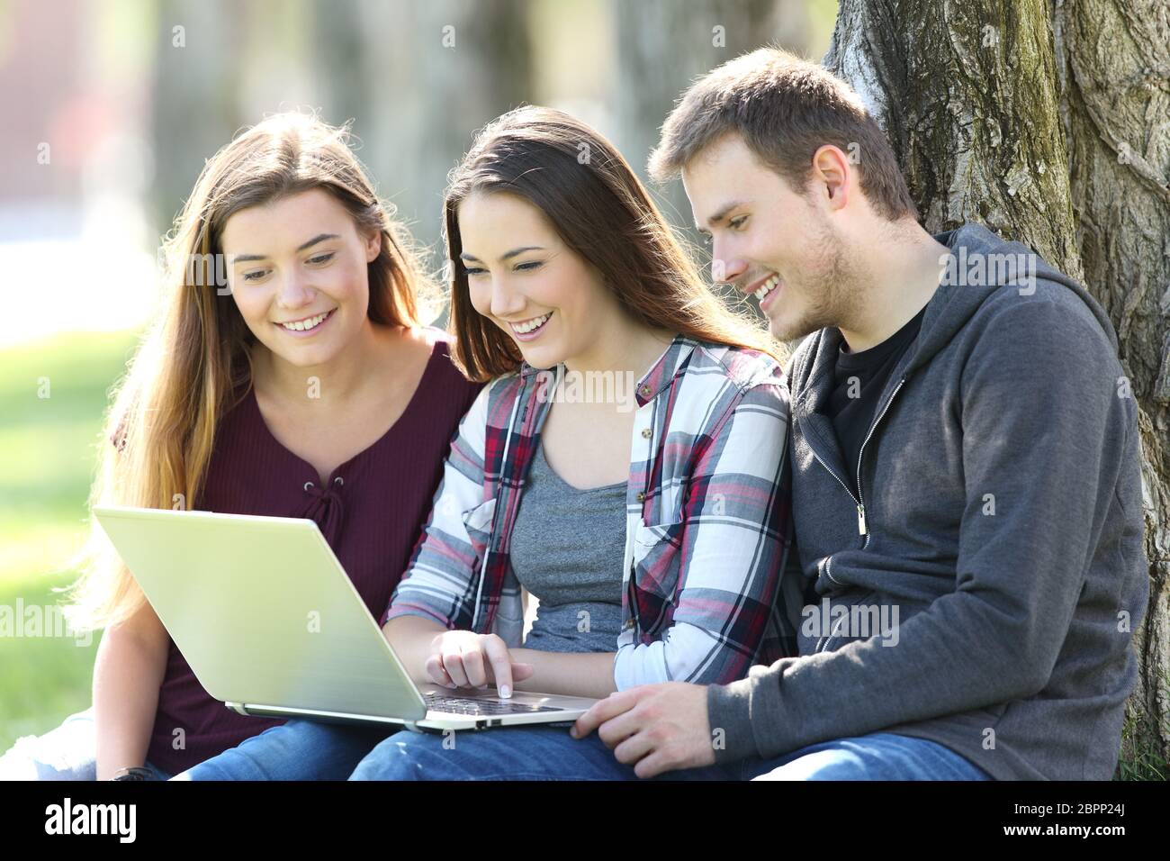 Three happy teenagers searching media content together on line with a laptop sitting on the grass in a park Stock Photo