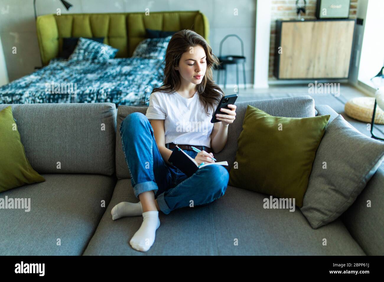 Close up of smiling woman sitting near sofa, writing in weekly planner and surfing phone. Working at home. Stock Photo