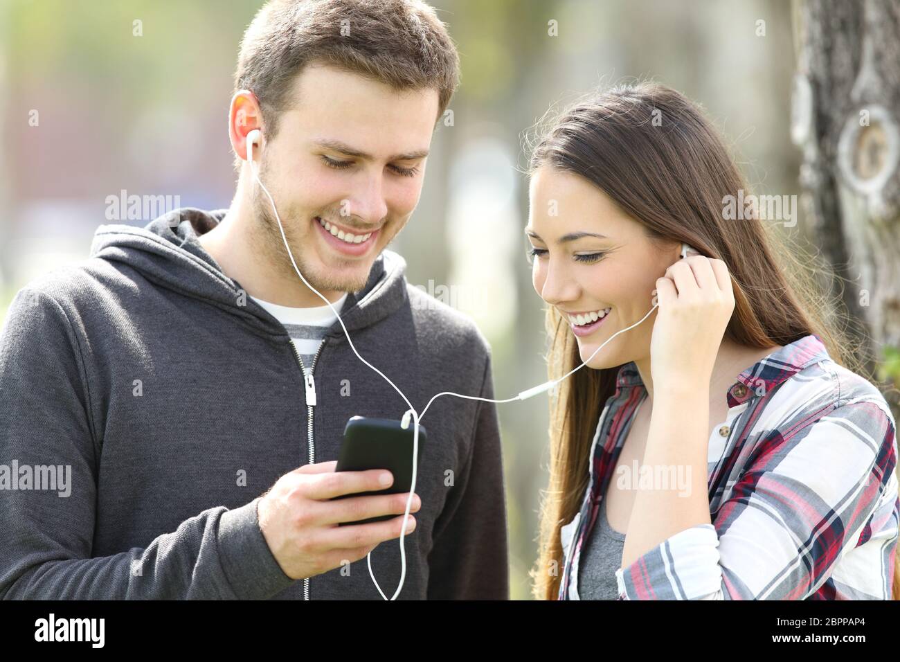 Happy couple of teenagers sharing on line music and watching media content in a smart phone outdoors in a park Stock Photo