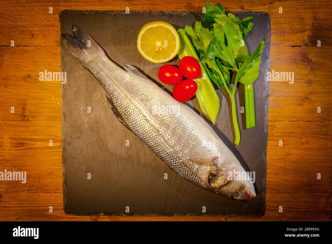 Slate dish with sea bass, lemon, celery and tomatoes on wooden background Stock Photo
