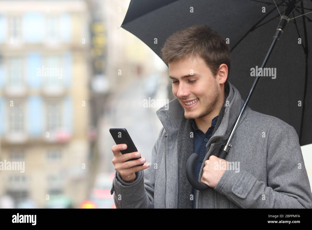 Happy man checking smart phone under an umbrella in winter in the street Stock Photo