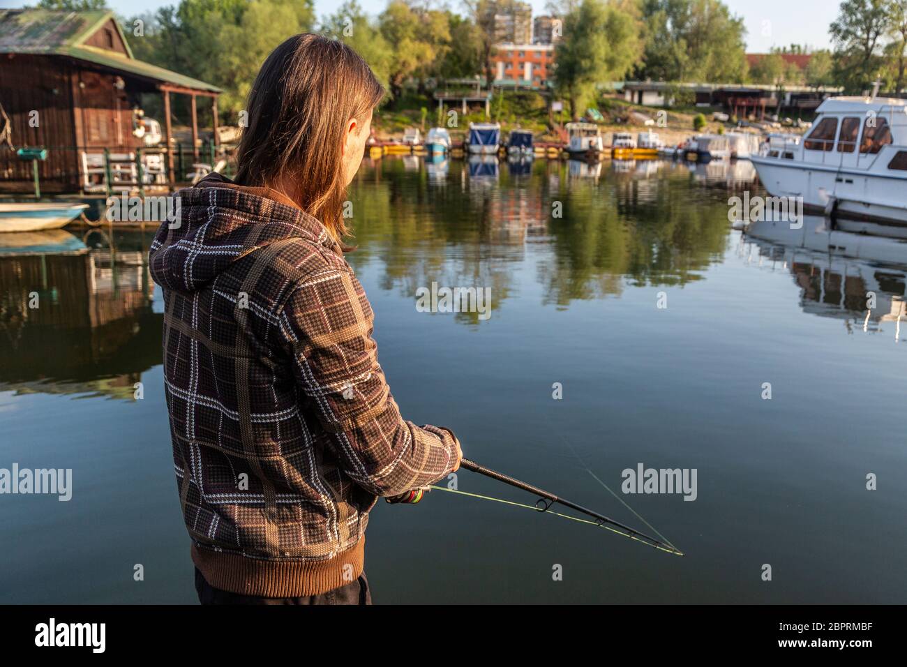 Man with long hair is fishing at river corner from raft. Boats and