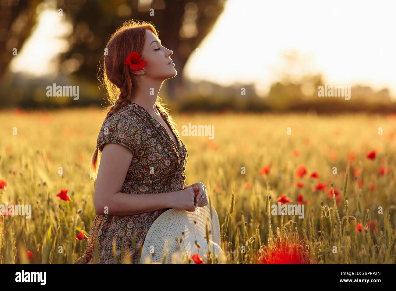 Redhead woman with hat on green field with poppies in summer sunset Stock Photo