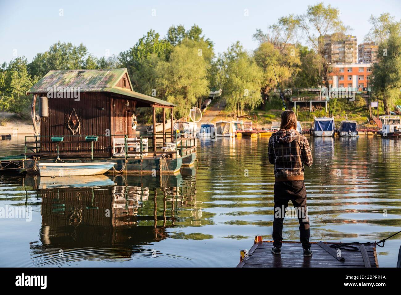 https://c8.alamy.com/comp/2BPRR1A/man-with-long-hair-is-fishing-at-river-corner-from-raft-boats-and-floating-houses-are-around-him-with-trees-in-background-he-is-using-fishing-rod-wi-2BPRR1A.jpg