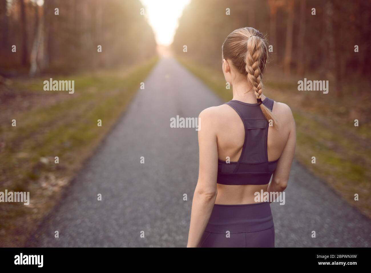 Sporty healthy young woman walking at sunrise along a rural road through a dense forest towards the glow of the sun at the end between the trees in a Stock Photo