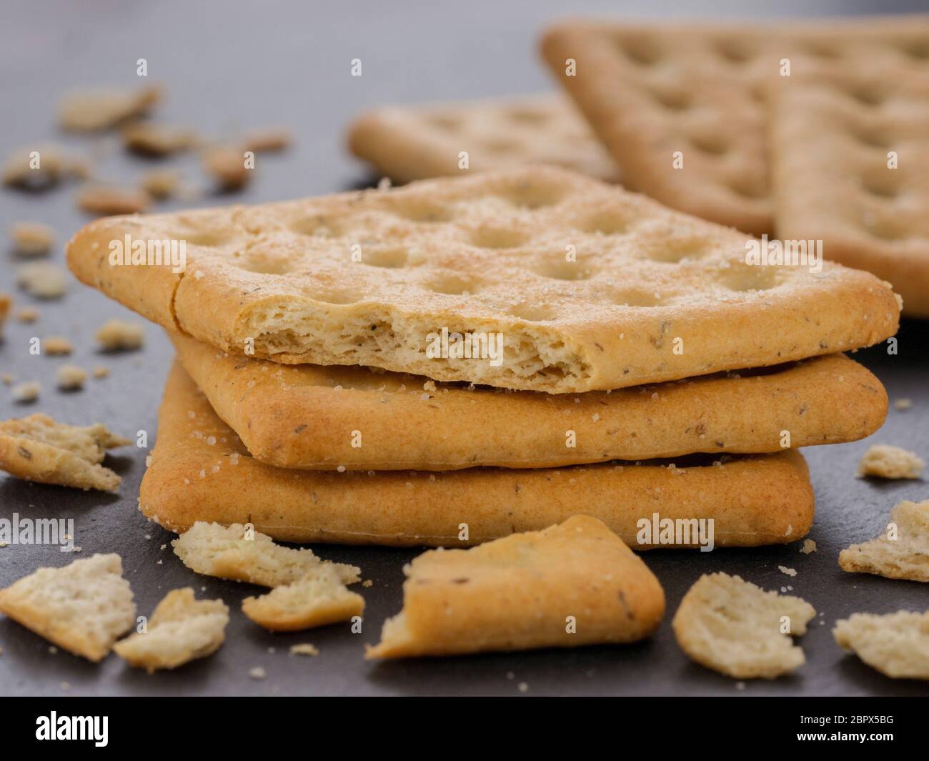 stack of square crackers with pieces and crumbs on slate gray background. Dry salt cracker cookies with fiber and dry spices Stock Photo