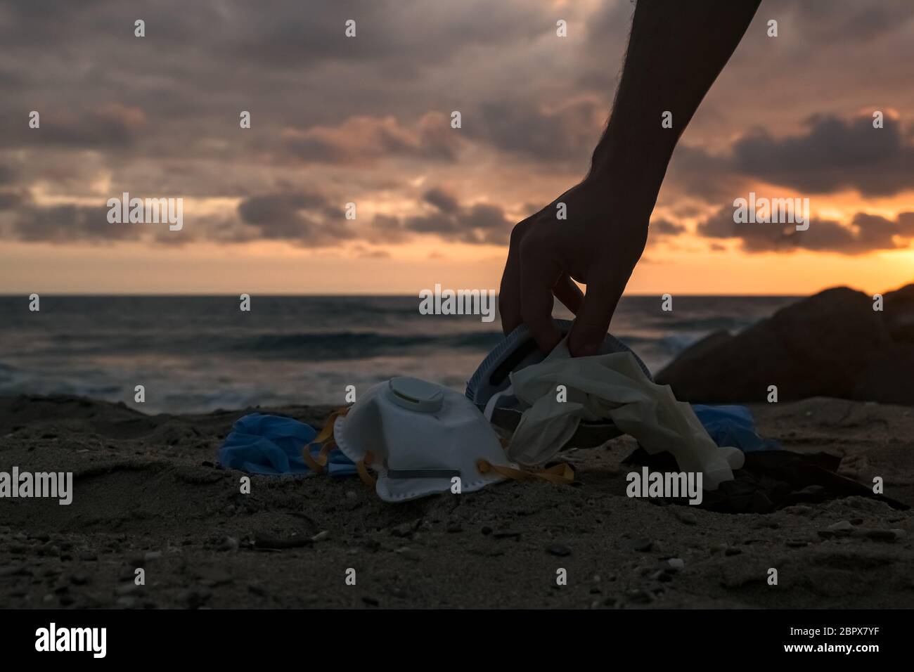 Man habd collect medical waste,mask and plastic gloves garbage trash on sunset sea shore,coronavirus covid pollution disease  Stock Photo