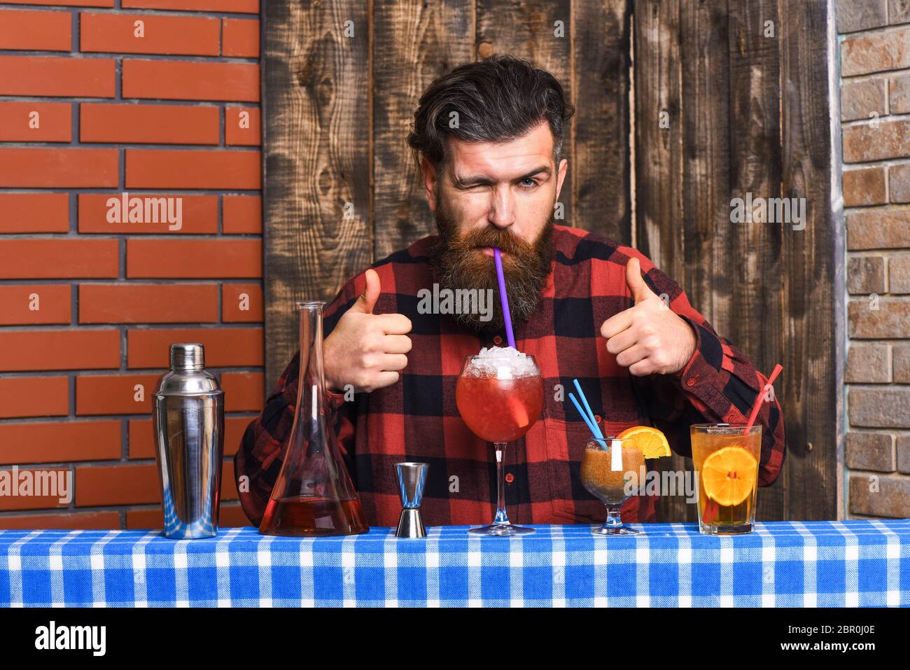 Hipster enjoy drink and thumbs up. Barman with beard and cheerful face drinks out of glass with drinking straw cocktail. Man in checkered shirt on wooden background. Barman and cocktails concept. Stock Photo