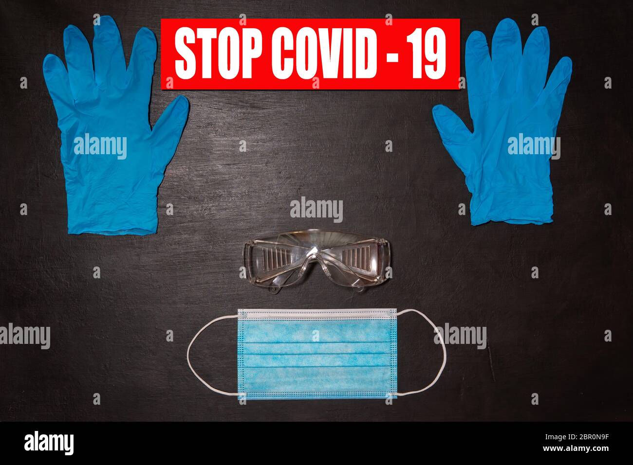 Black medical background. Safety goggles, medical mask and two blue gloves. On the gloves there is a banner with the words Stop Covid-19 Stock Photo