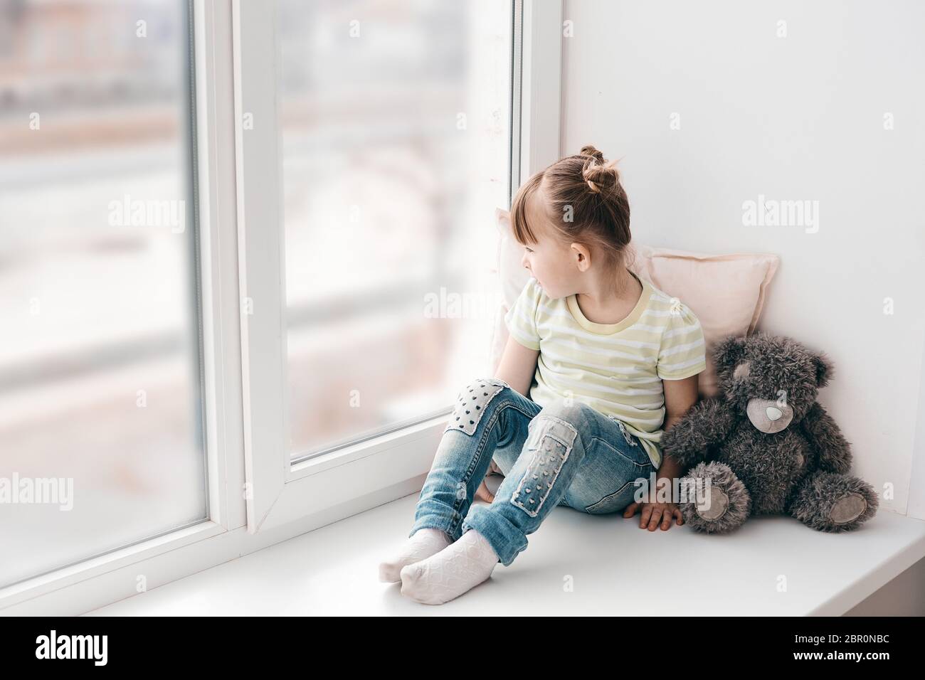 A little girl sits on a window sill and looks out the window into the street. It's a sad mood. Illness or self-isolation. Stock Photo