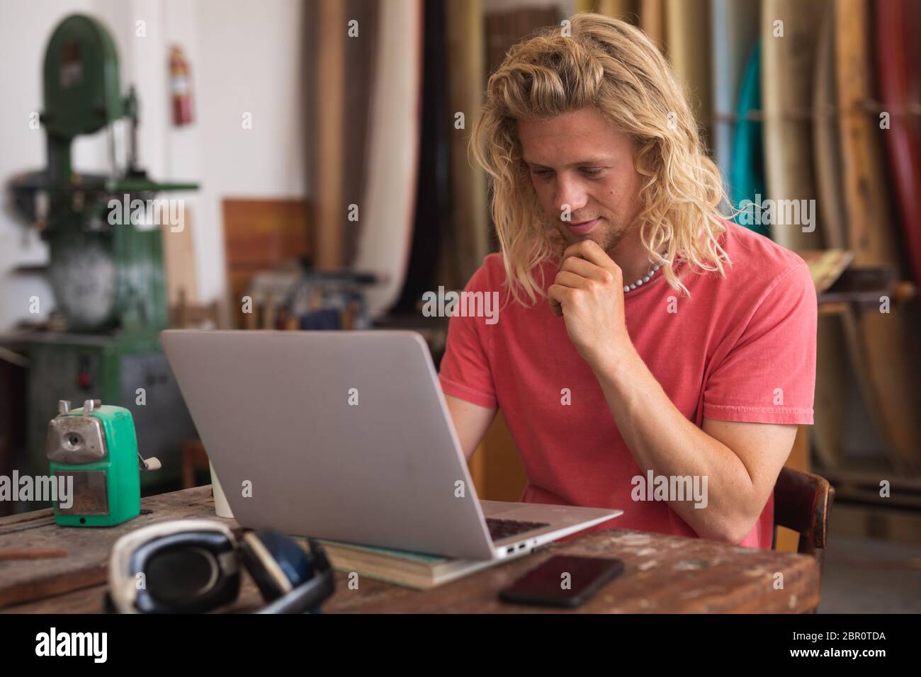 Caucasian male surfboard maker sitting at desk and using his laptop computer with surfboard behind Stock Photo