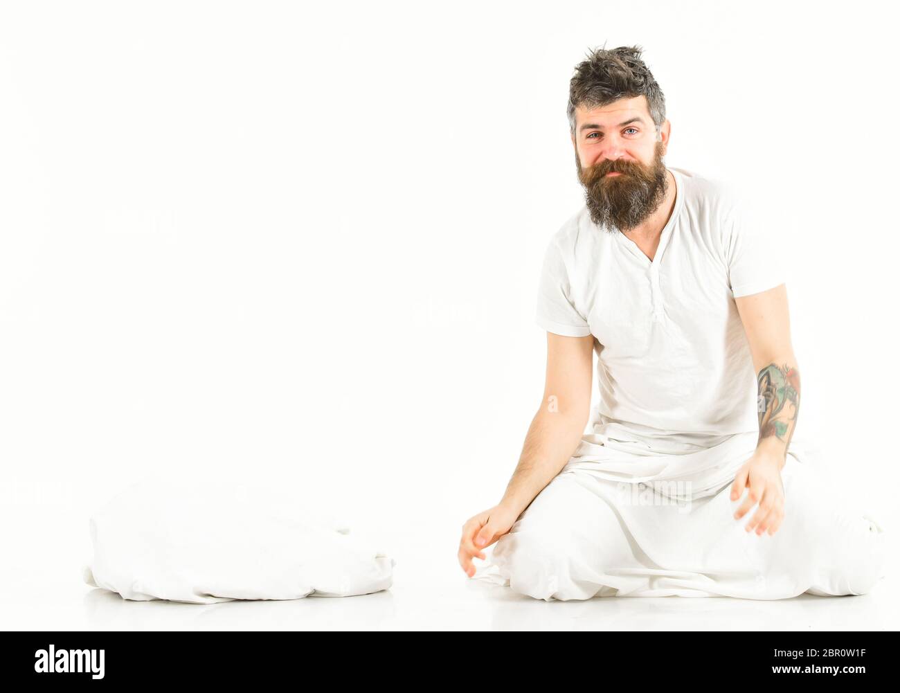 Hipster with beard sits near pillow, sleepy smiling macho. Man after good rest in morning, white background. Energetic man concept. Man with happy face in white pajama waking up. Stock Photo