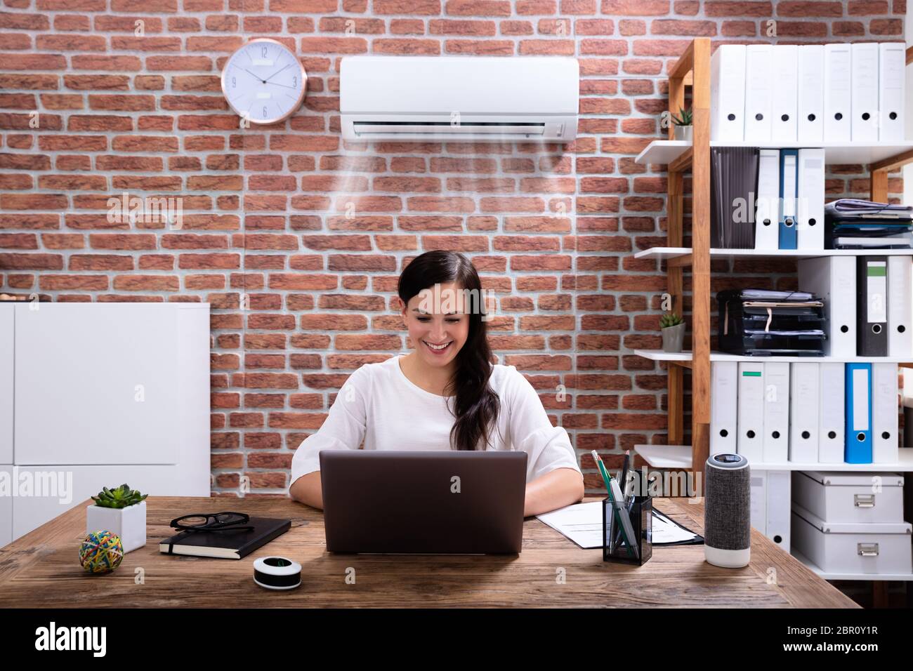 Smiling Businesswoman Enjoying The Cooling Of Air Conditioner Using Laptop At Workplace Stock Photo