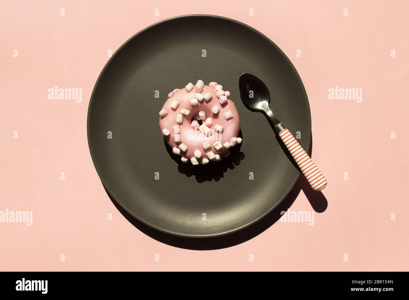 Pink donat decorated with white marshmallows on the grey ceramic plate near small stripped spoon on light coral background. Top view flat lay. Trendy Stock Photo
