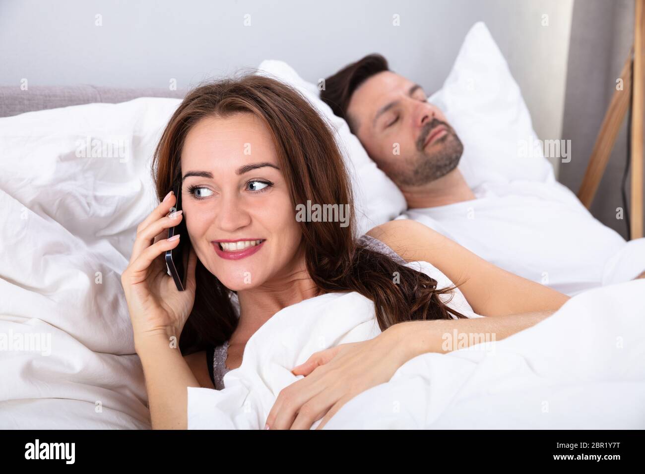 Smiling Young Woman Talking On Smartphone Besides Her Husband Sleeping