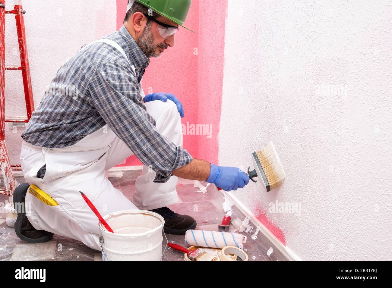 Caucasian house painter worker in white overalls, with helmet and goggles painting the pink wall with white paint using the brush. Construction indust Stock Photo