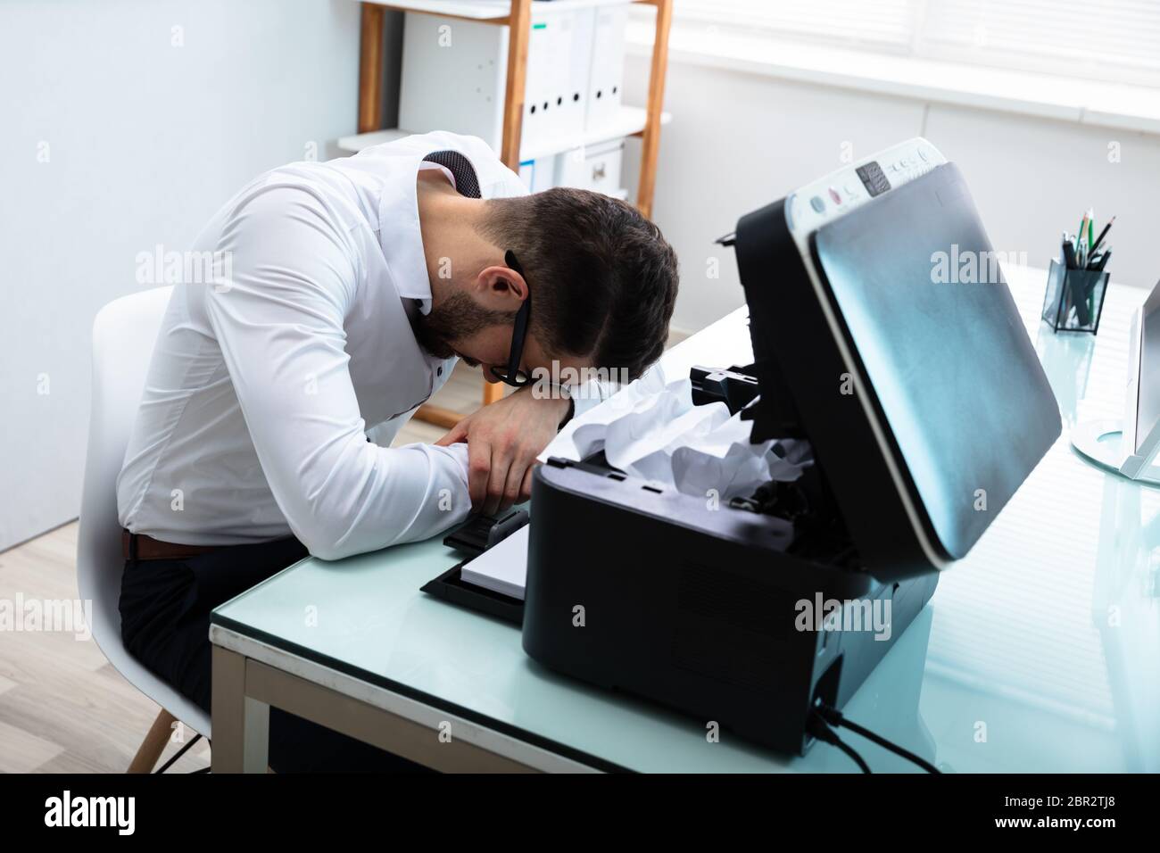 Tired Businessman Resting His Head In Front Of Paper Stuck In Printer Stock Photo