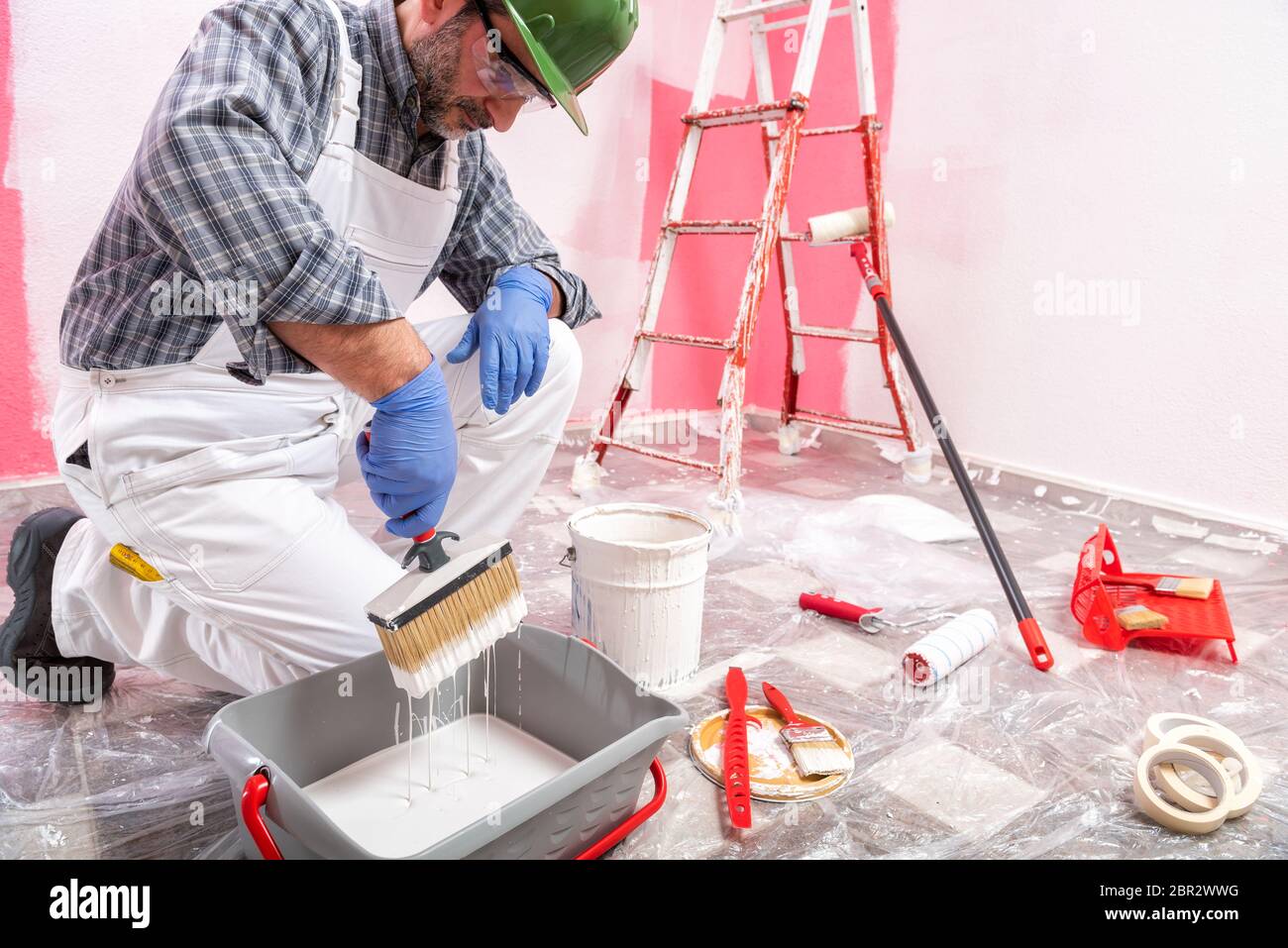 Caucasian house painter worker in white overalls, with helmet and goggles he prepares the white paint to paint the pink wall. Construction industry. W Stock Photo