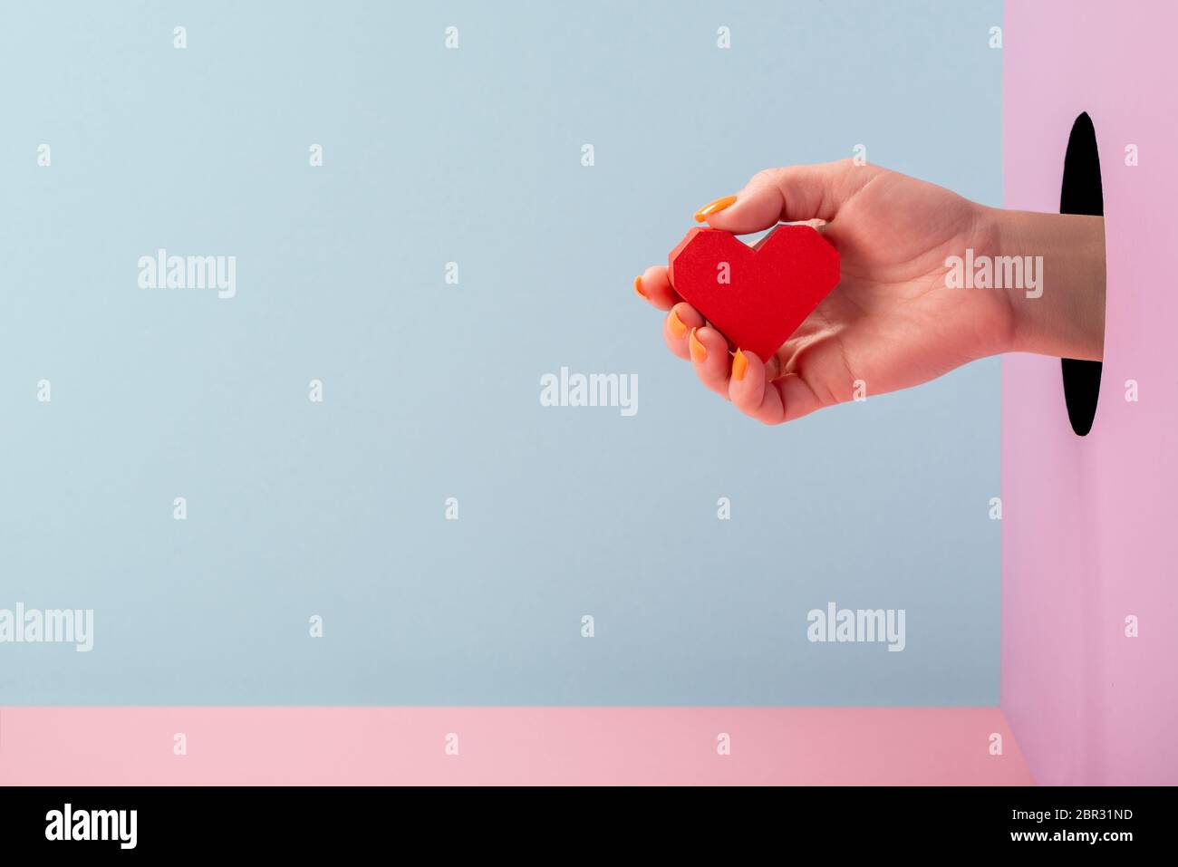 Woman's hand holding a red paper heart on blue and pink background ...