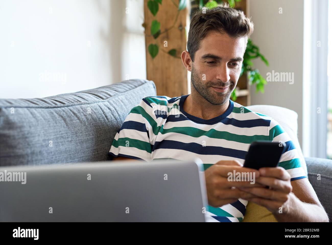 Young man using a cellphone and laptop on his sofa Stock Photo