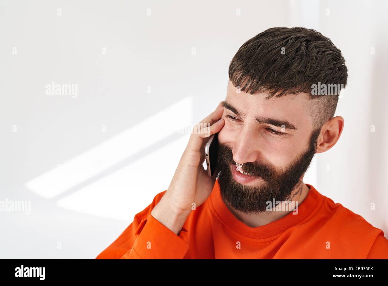 Image of young bearded man with nose jewelry wearing orange shirt talking on smartphone while standing over white wall indoors Stock Photo