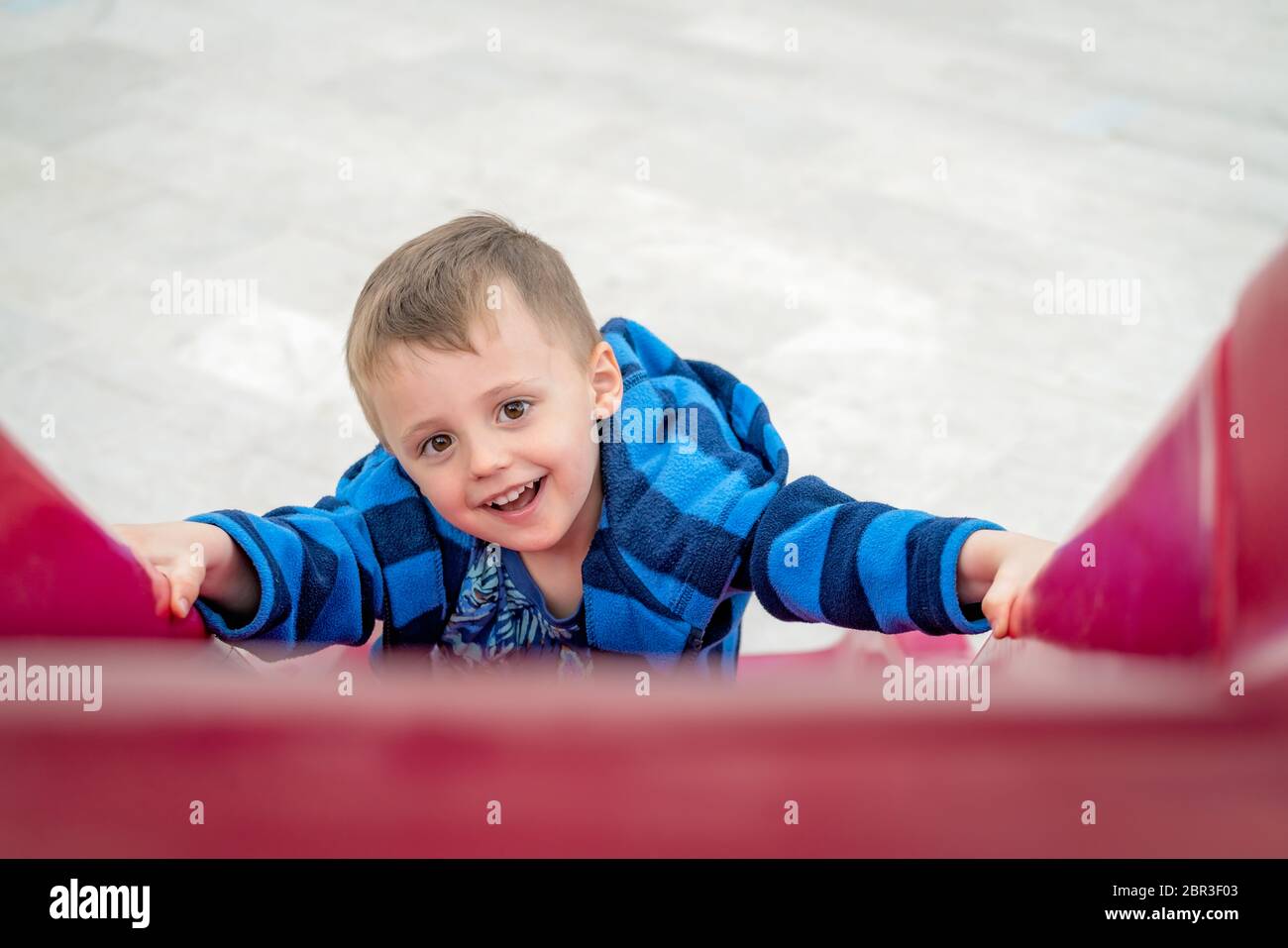 Little cheeky mischevious Caucasian boy climbing up the red plastic outdoor slide on a playground Stock Photo