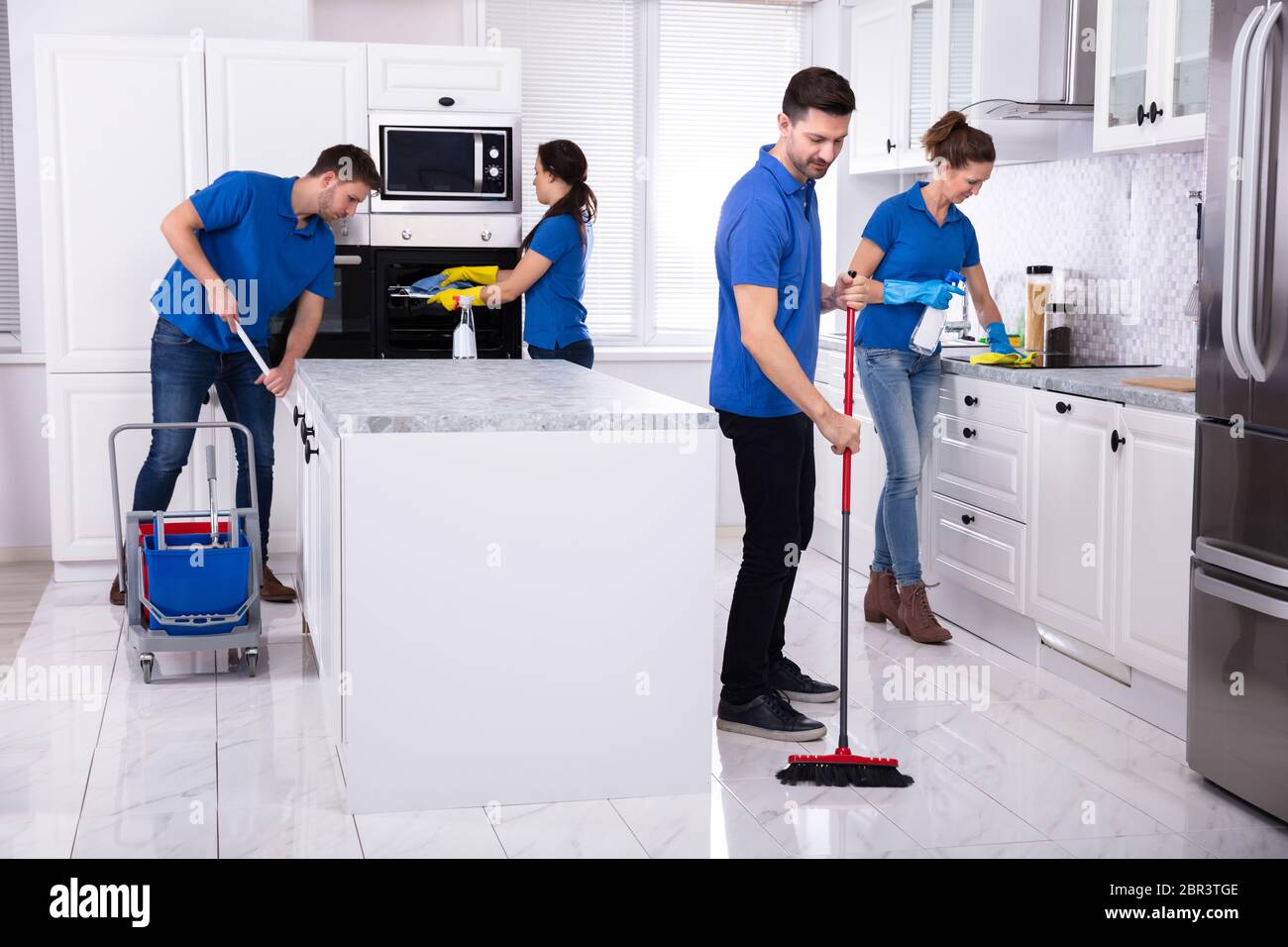Group Of Young Janitors In Uniform Cleaning Kitchen At Home Stock Photo
