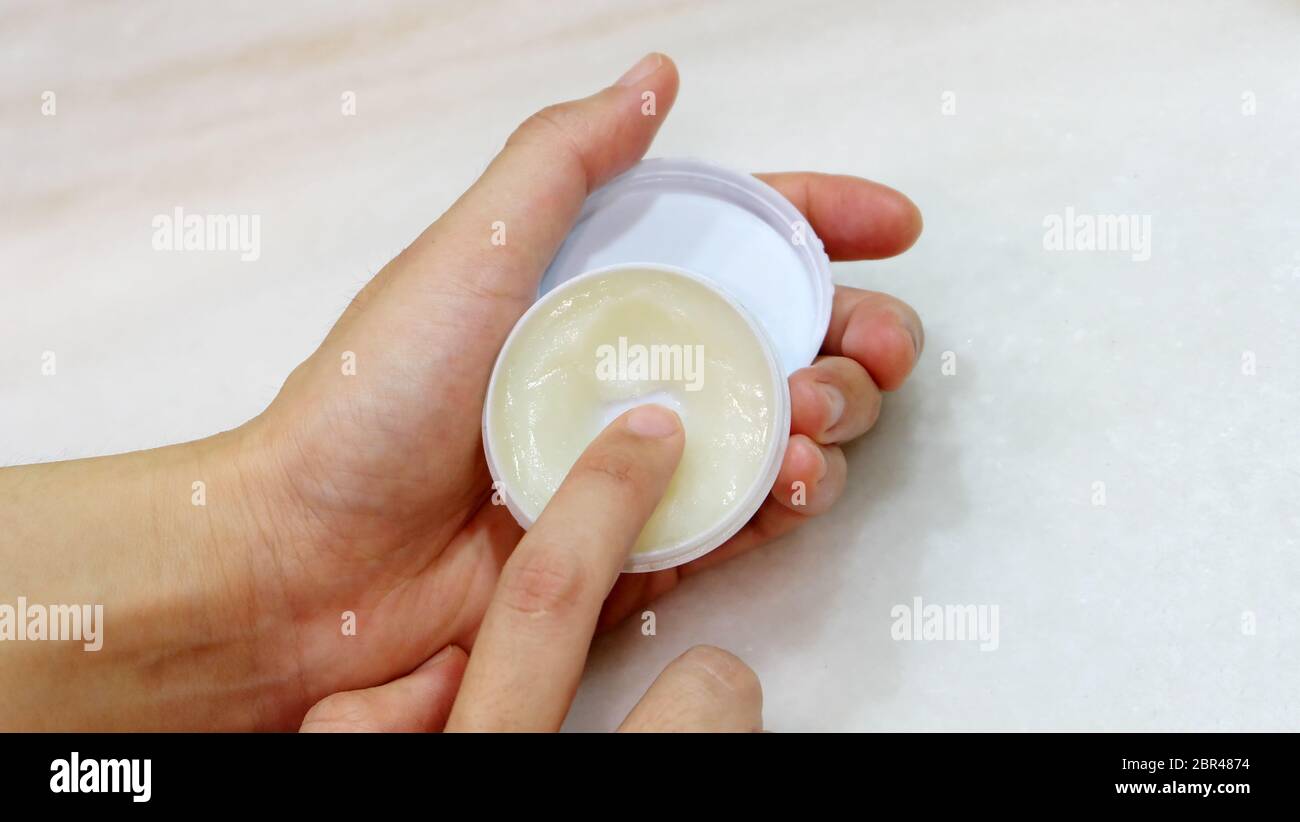Hand holding a small round container with ointment, and a finger trying to scoop some ointment out. Stock Photo