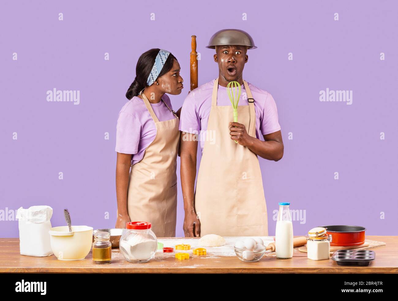 Angry black girl with rolling pin and her frightened boyfriend trying to bake together on color background Stock Photo