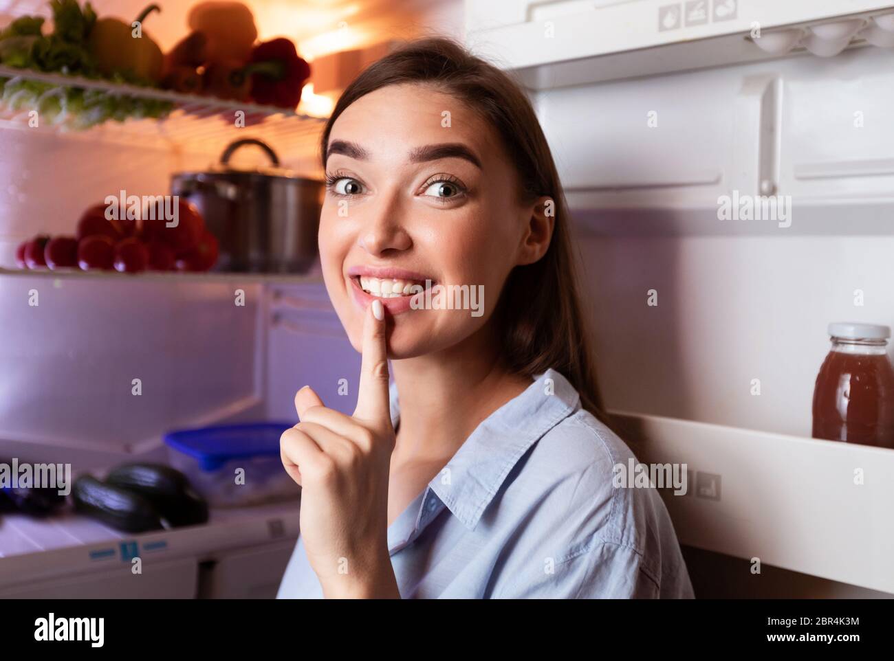 Tricky girl putting finger on lips and smiling near fridge Stock Photo