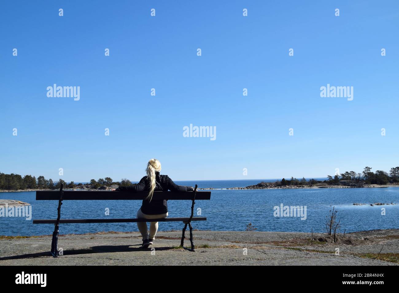 Blonde young woman, in her 30s, all alone, sitting on a bench, looking out to sea and the archipelago in Sweden on a sunny day. Stock Photo