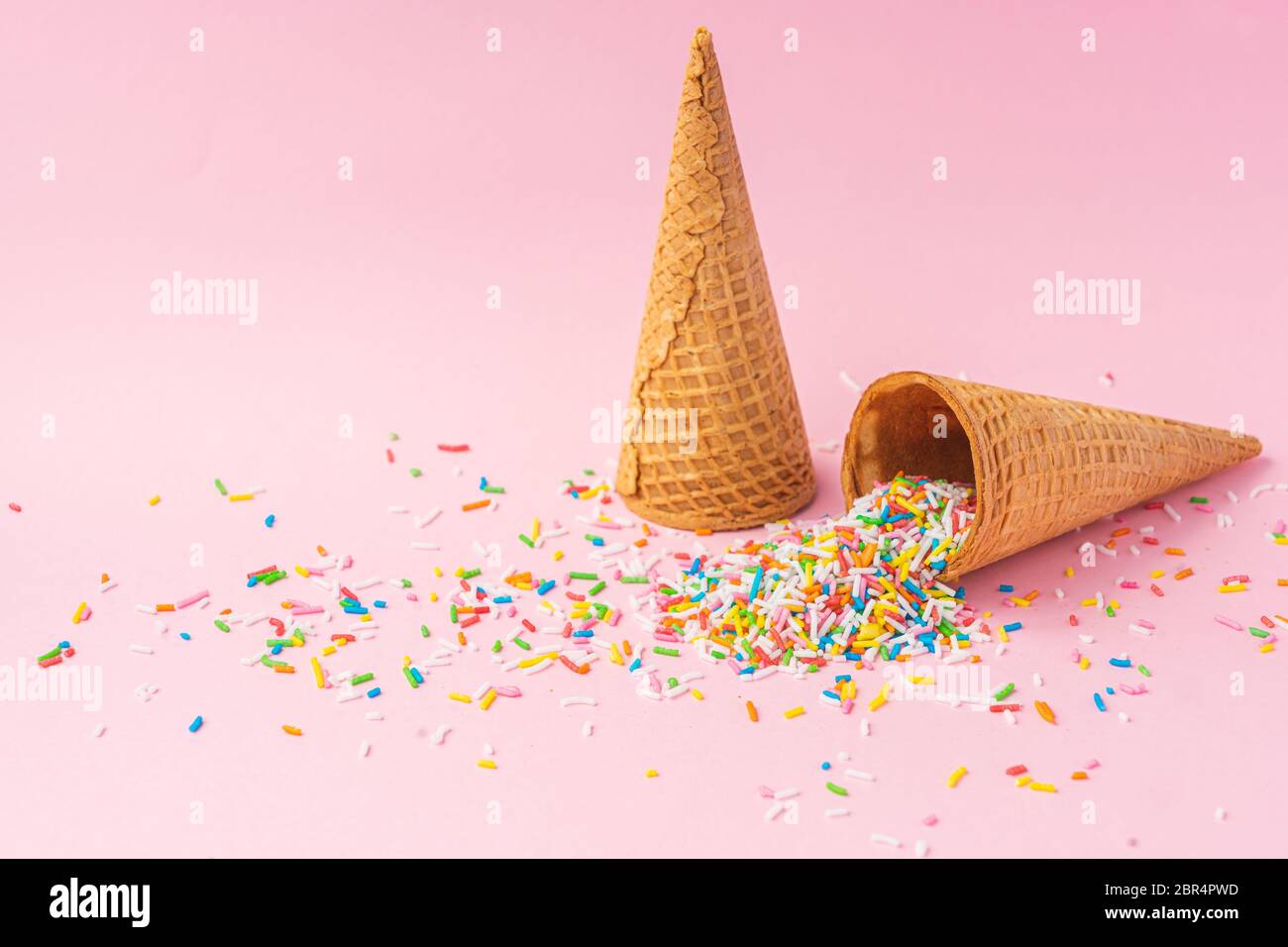 Two wheat flour ice cream cones one upside down and the other filled with colored sugar on a pink background. Summer concept. Stock Photo