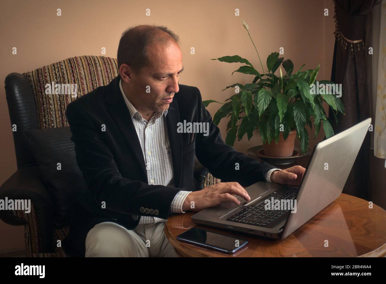 Smart clothed intelligent and professional senior adult man teleworking from home writes essay from his laptop while sitting on his armchair with plan Stock Photo