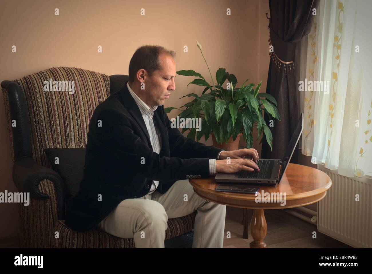 Smart clothed intelligent professional senior adult man teleworking from home writes essay from his laptop while sitting on his armchair with plant on Stock Photo