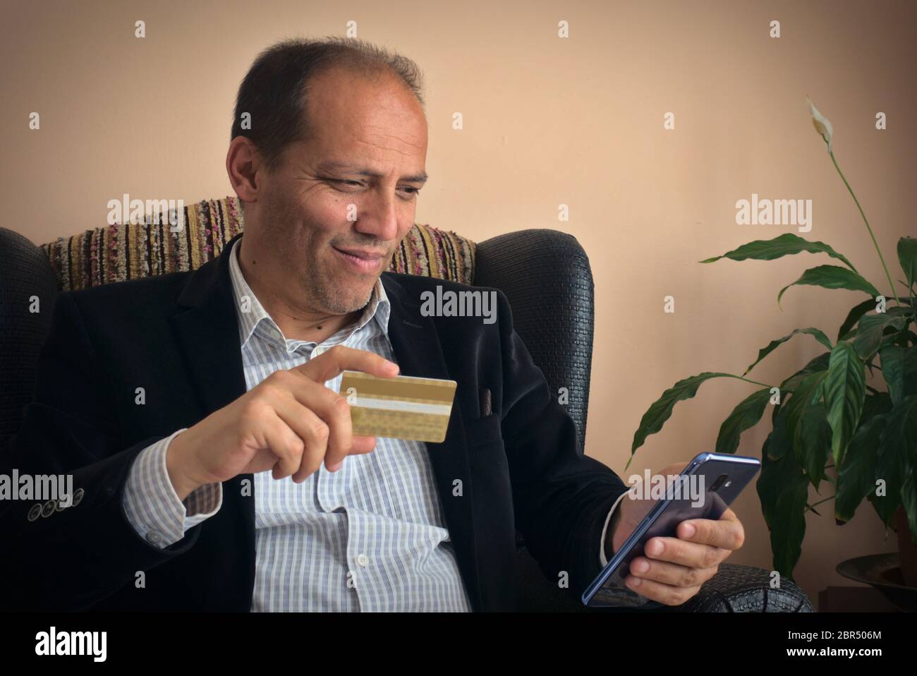 Intelligent, happy and smiling senior adult male in smart clothes holding a credit card shopping online by using his smart phone, with a plant on back Stock Photo