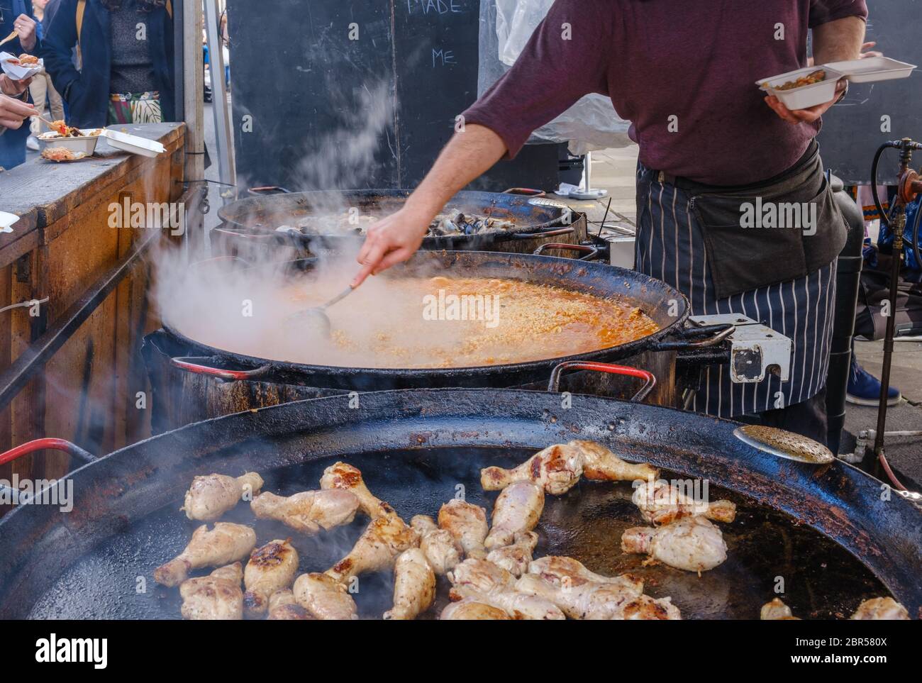 A man serves paella into a container next to large pans of cooking