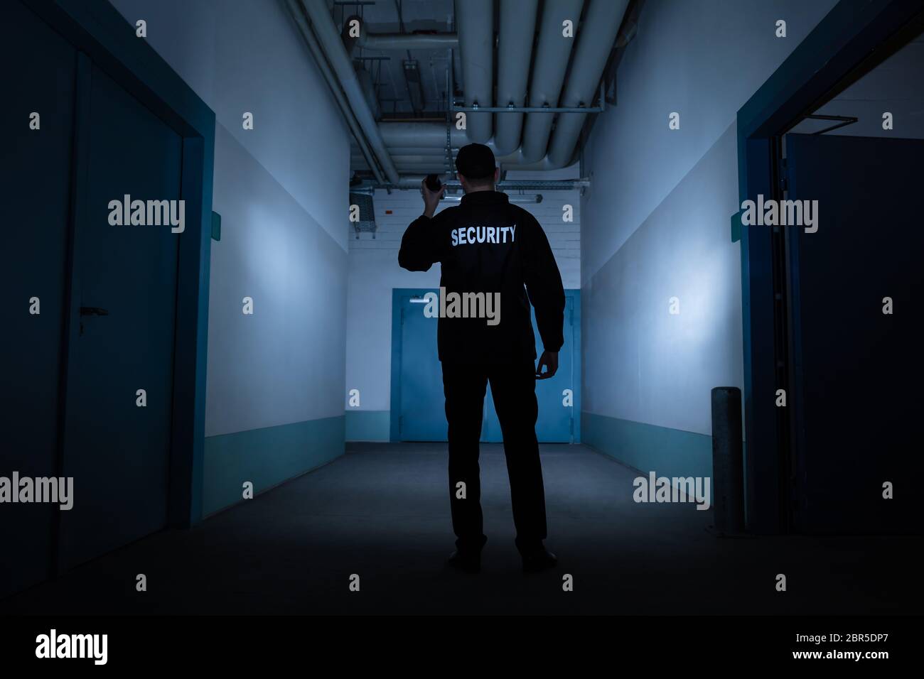 Rear View Of A Male Security Guard With Flashlight Standing In Corridor Stock Photo