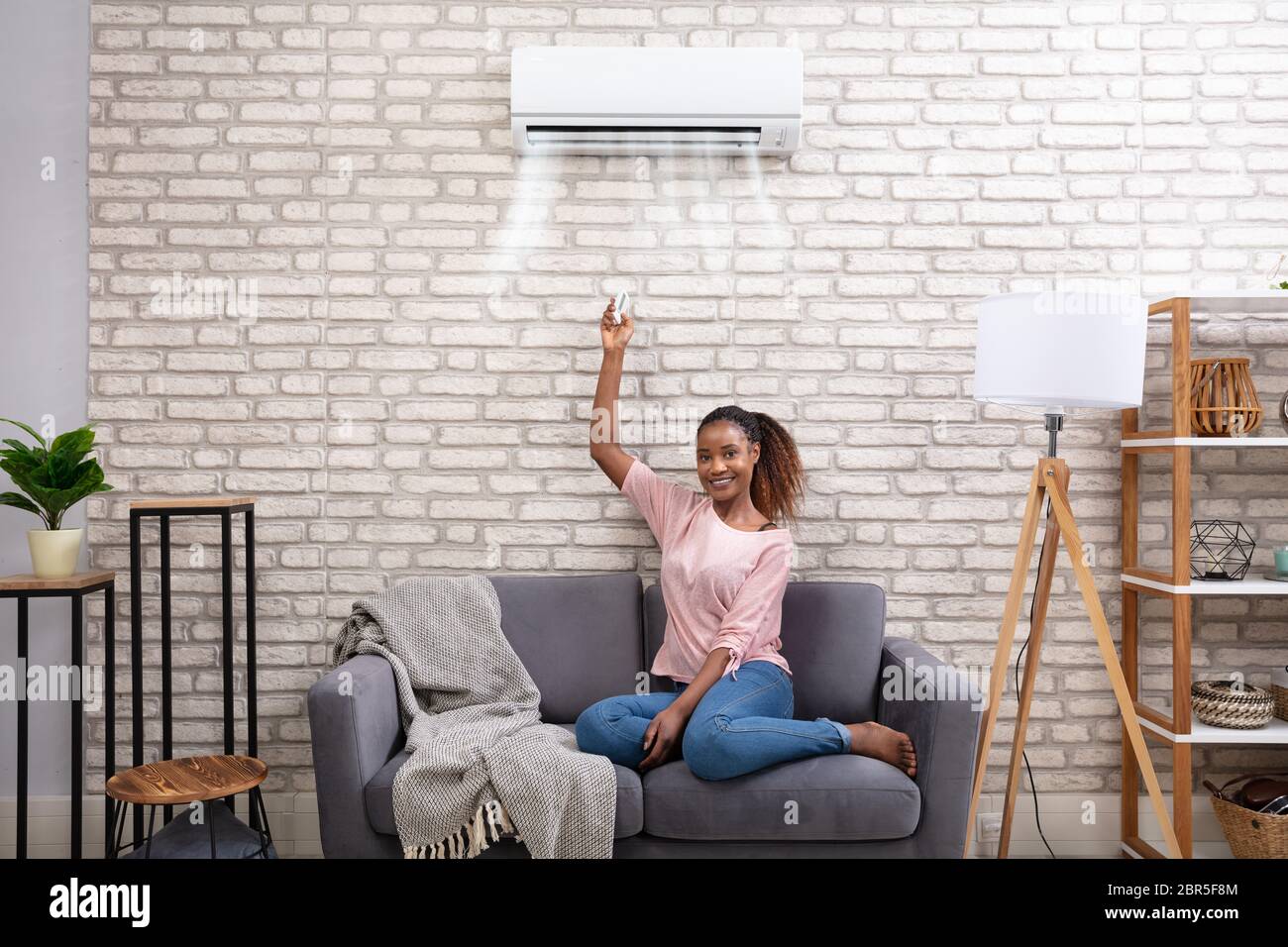 Young African Woman Switching On Air Conditioner With Remote Control At Home Stock Photo