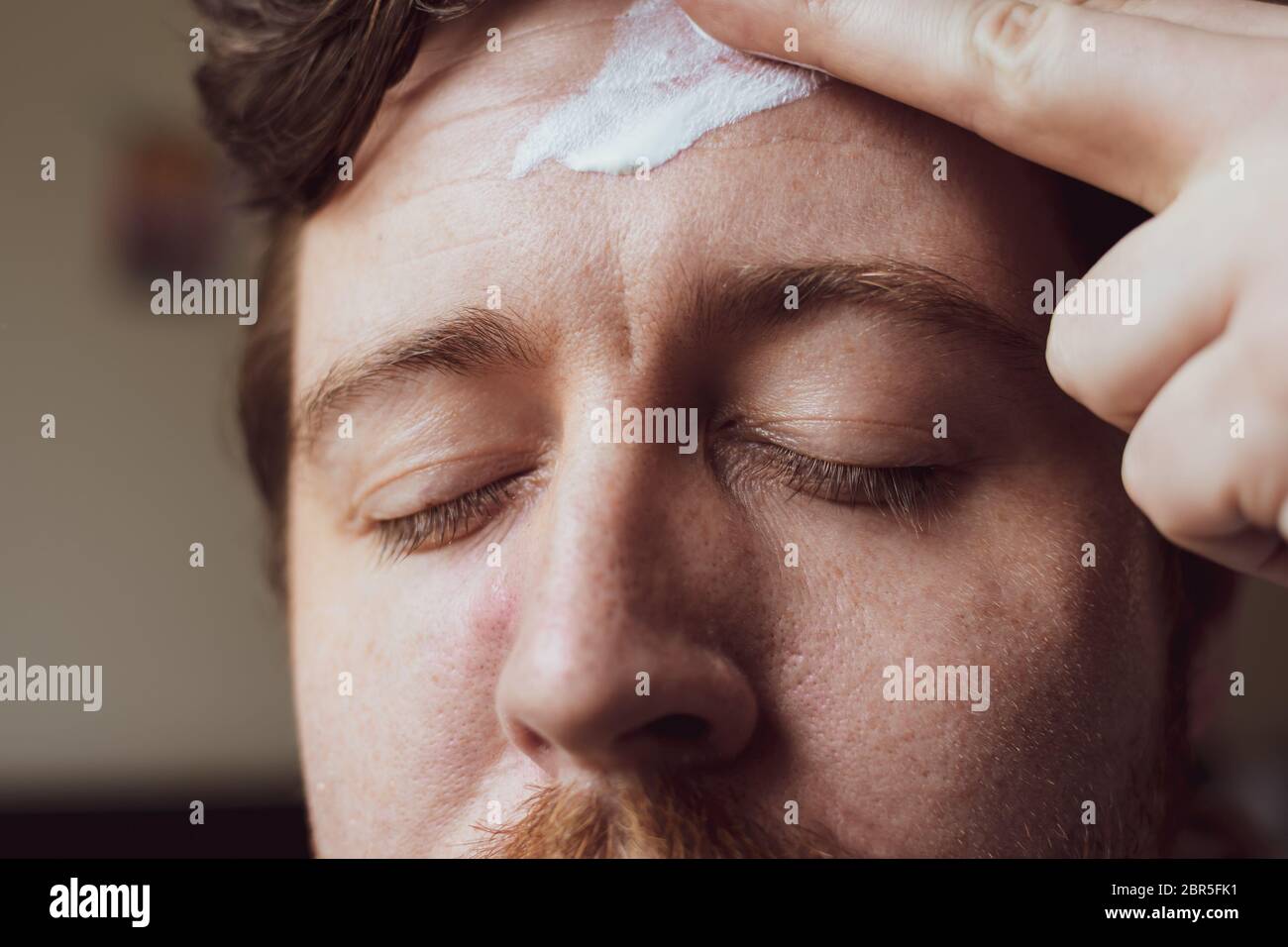 A close up of the face of a man in his thirties as he uses face cream to moisturise his forehead as part of a skincare routine Stock Photo