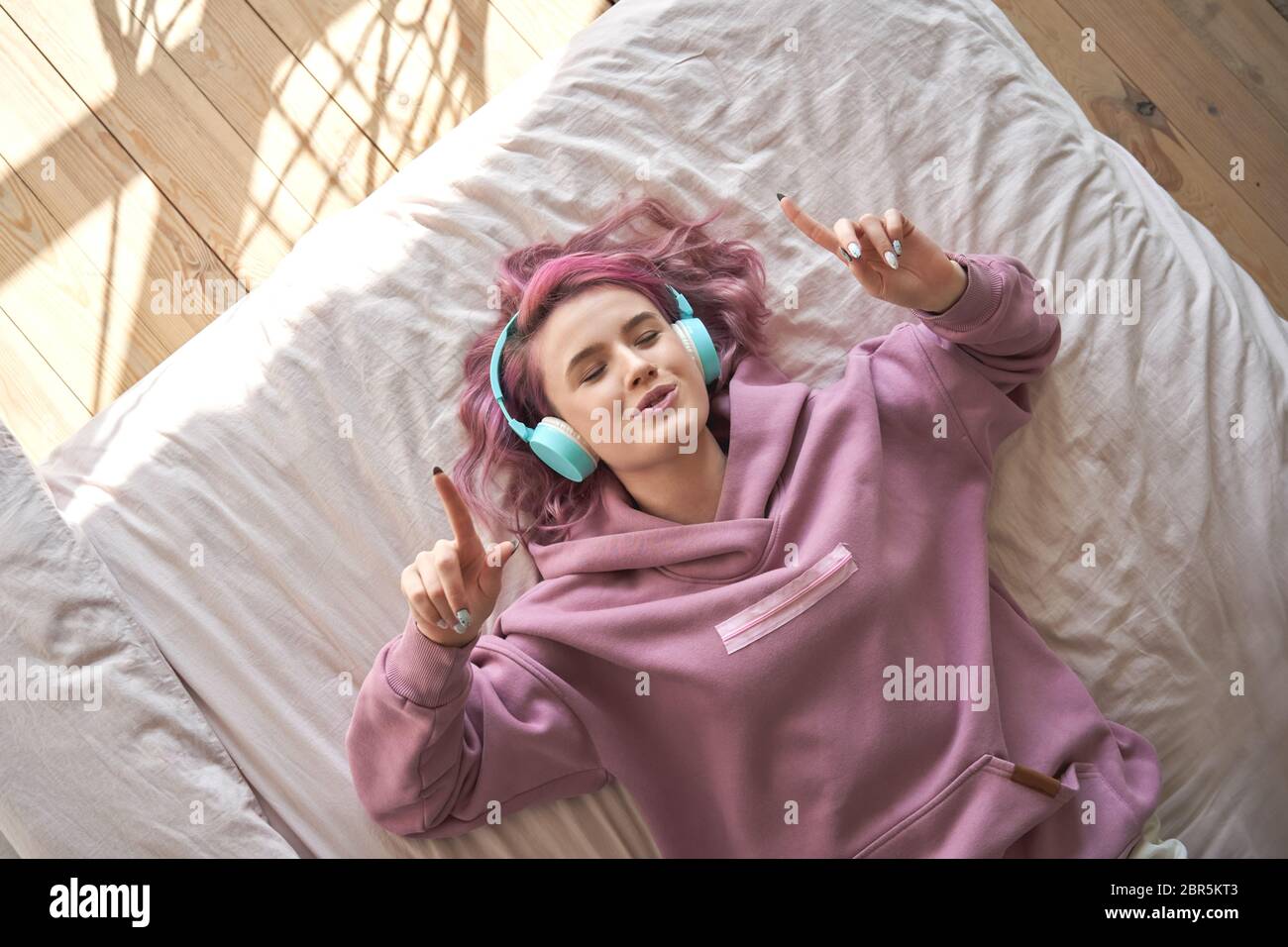 Happy teen girl pink hair wear headphones lying in bed listening music