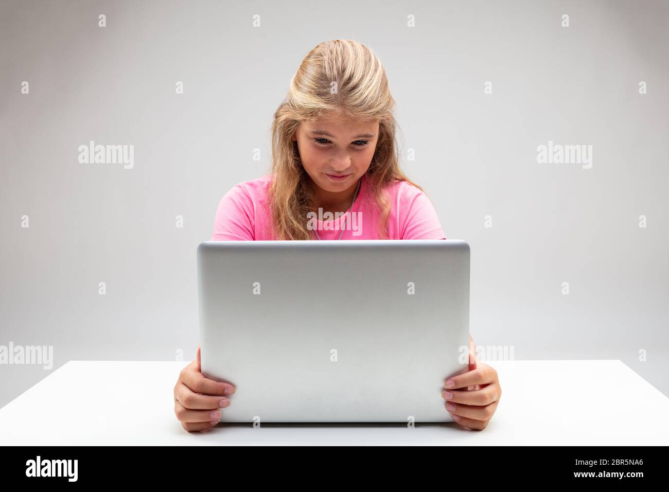 Young blond girl looking at her laptop with distaste grimacing in revulsion as she holds the screen with both hands seated at a small white table Stock Photo