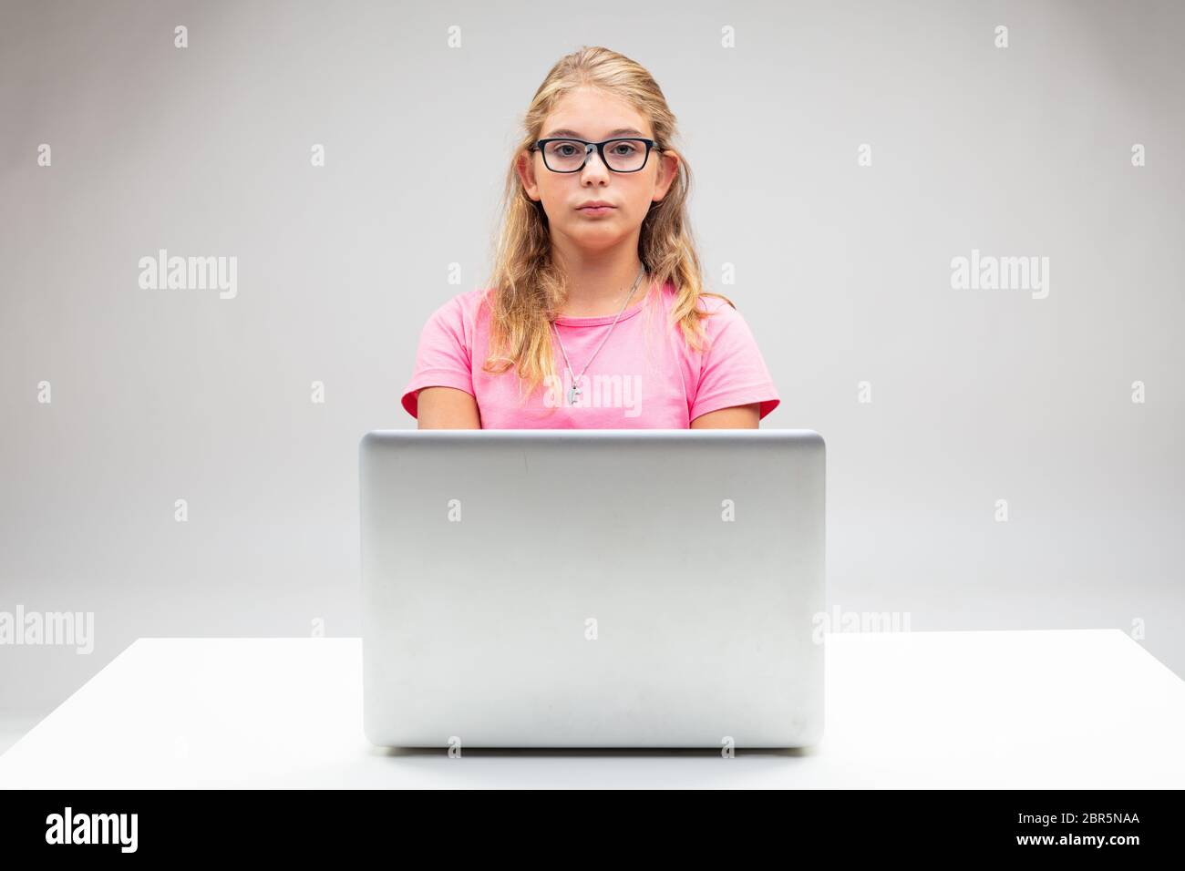Proud little blond girl seated at her laptop with a deadpan stern expression wearing glasses in a spoof of a scholarly student or businesswoman Stock Photo