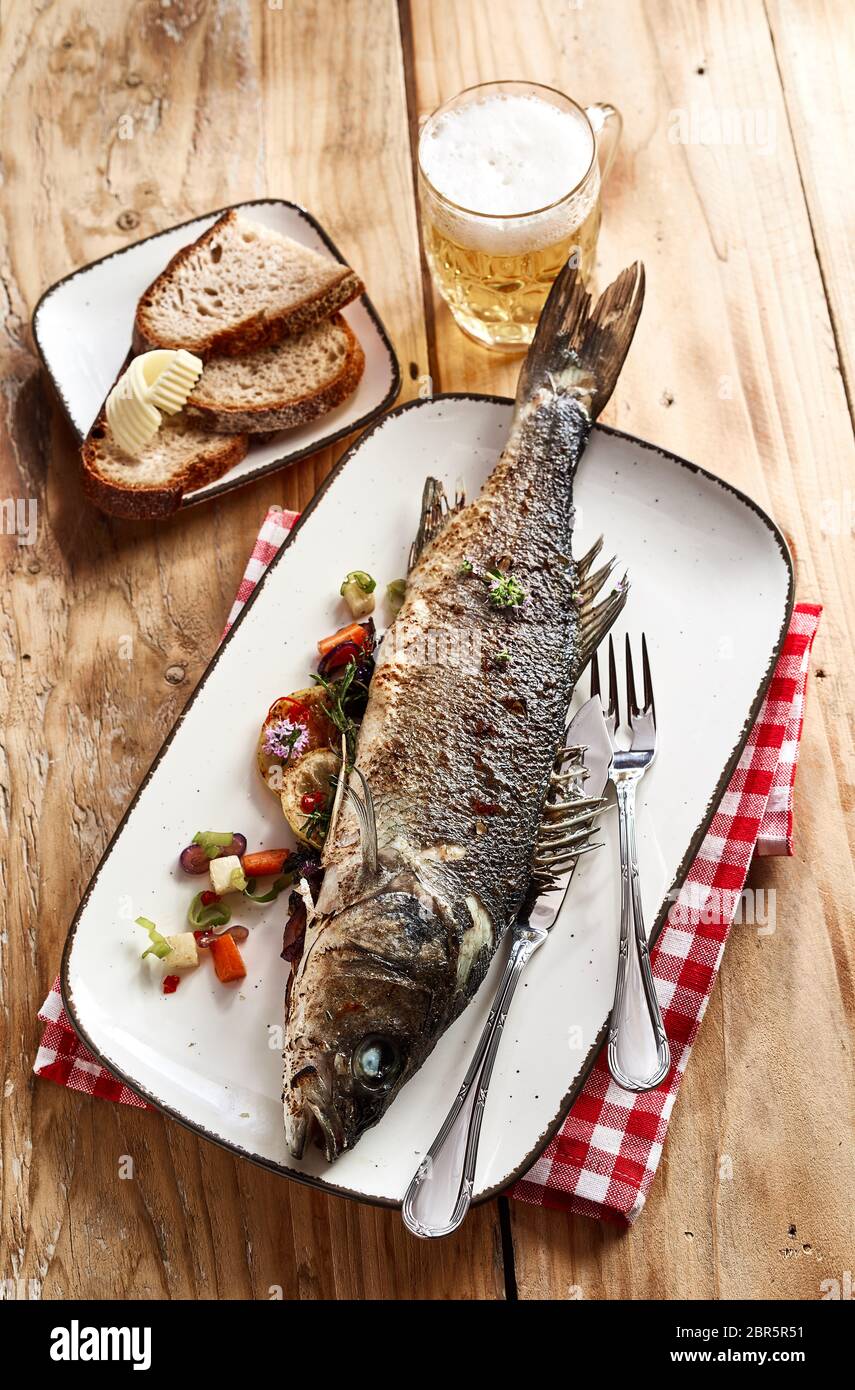 Whole oven grilled sea bass with a cold beer served with rye bread on a rustic or vintage wood