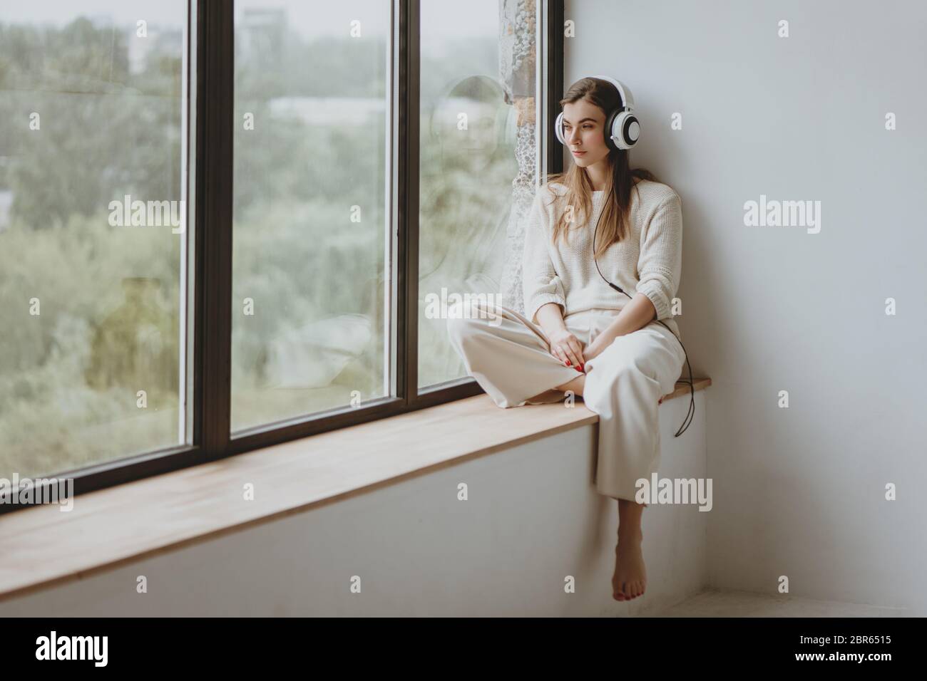 Young beautiful woman sitting on windowsill and listening music in headphones. Stock Photo