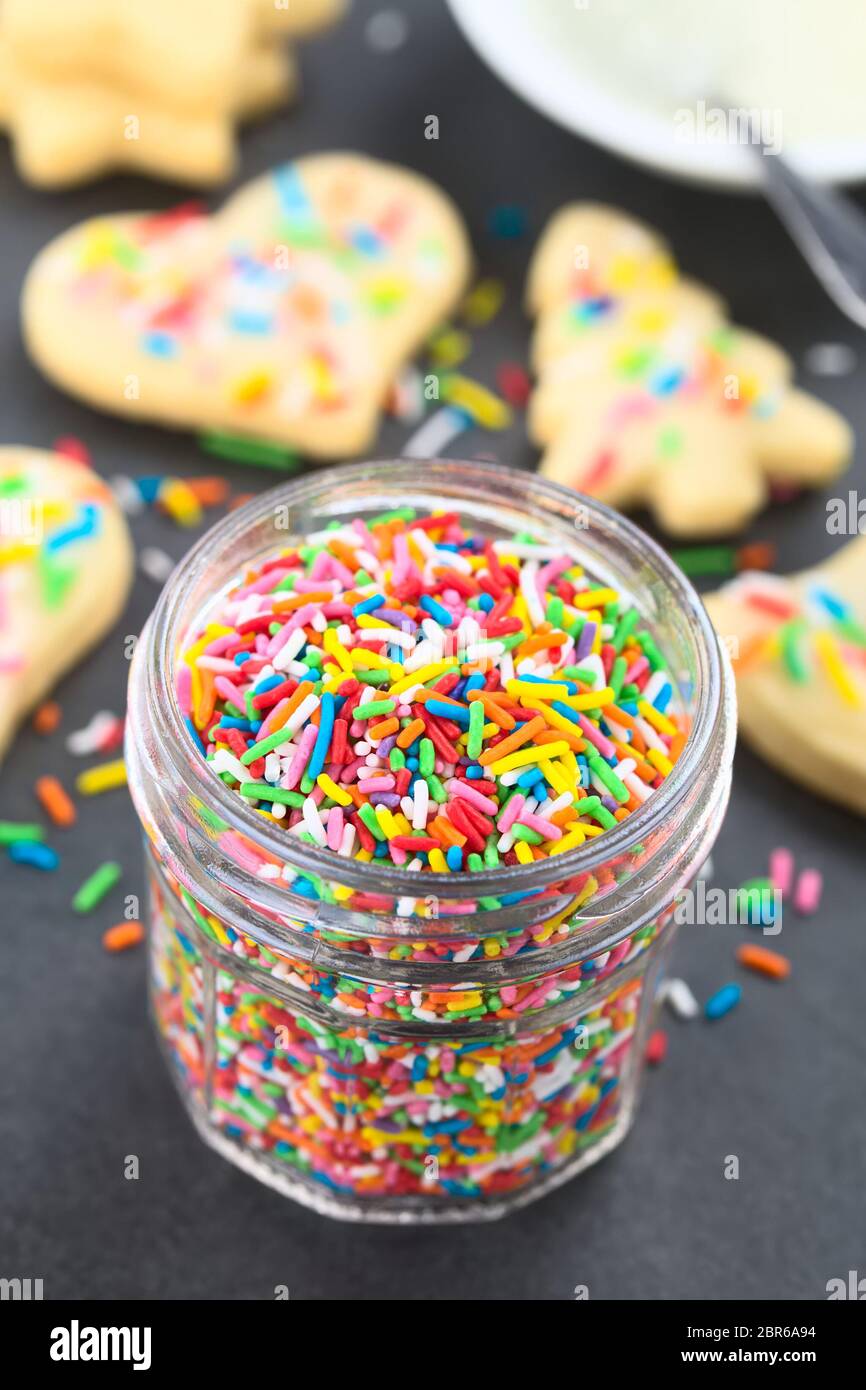 Colorful sugar sprinkles in glass jar with decorated sugar cookies and icing in a bowl in the back, photographed on slate (Selective Focus, Focus one Stock Photo