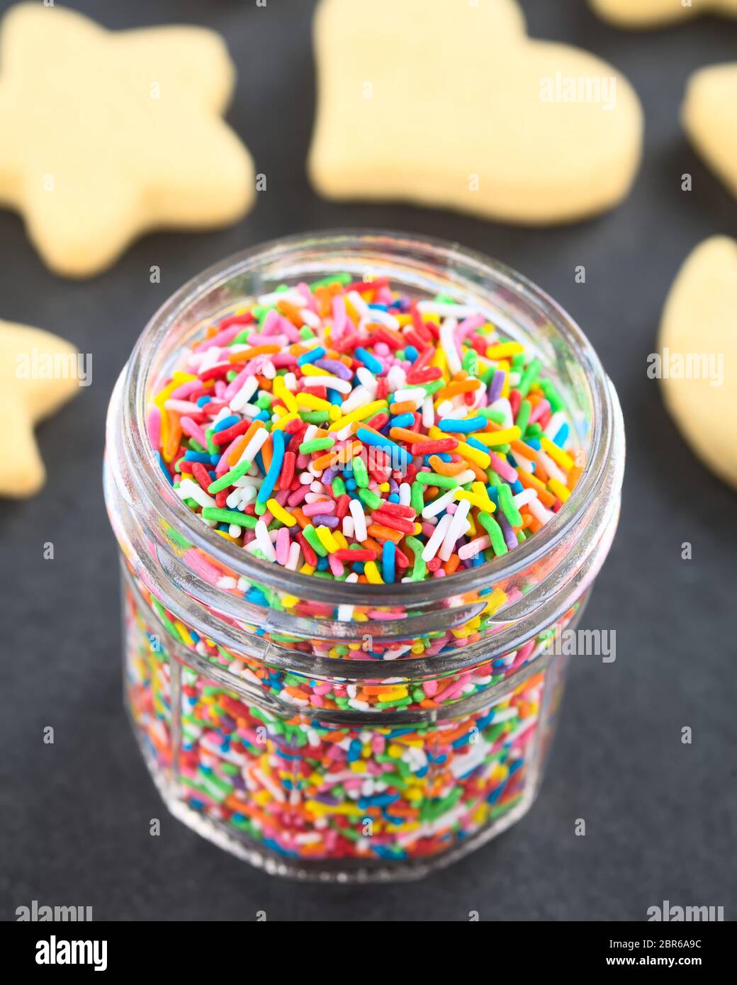 Colorful sugar sprinkles in glass jar with undecorated sugar cookies in the back, photographed on slate (Selective Focus, Focus one third into the spr Stock Photo