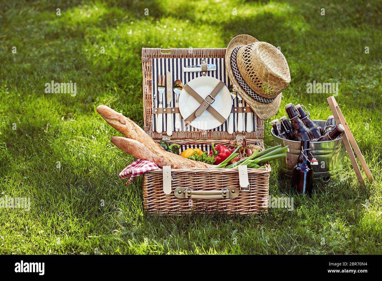 Vintage wicker picnic hamper filled with fresh vegetarian food and French baguettes beside a silver wine cooler with beers on green summer grass Stock Photo